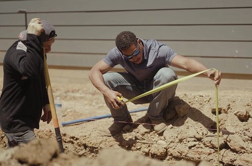 Corey crouching in a footing trench, measuring with a tape while a crew member holds a shovel
